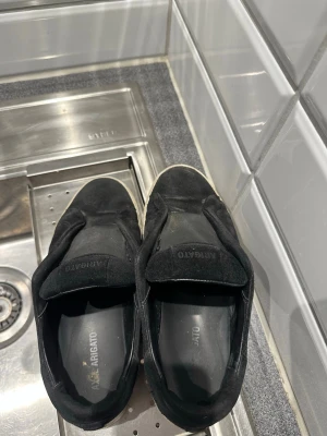 Svarta slip-on sneakers från Axel Arigato - Svarta slip-on sneakers från Axel Arigato med ovandel i mocka och diskret logga på tungan. Skorna har en vit, tjock gummisula och minimalistisk design utan snörning. Perfekta för en clean och stilren look.