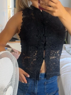 Svart spetsblus med knäppning - Säljer en svart ärmlös blus i spets med dekorativa knappar framtill. Blusen har hög hals och vackra blommiga spetsdetaljer som ger en elegant look. Perfekt att styla med jeans eller kjol för en trendig outfit.