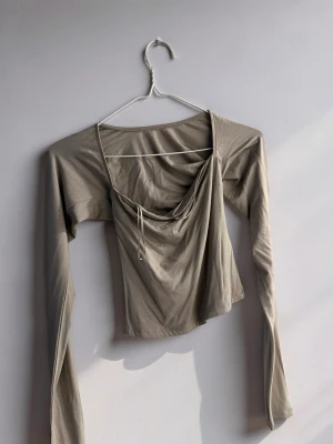 Beige långärmad topp med snörning - Trendig beige långärmad topp med fyrkantig ringning och snygg snörningsdetalj framtill. Toppen har en croppad passform och är tillverkad i ett mjukt, stretchigt material som känns skönt mot huden. Perfekt för dig som gillar stilrena och moderna plagg. (PRIS LAPP KVAR)