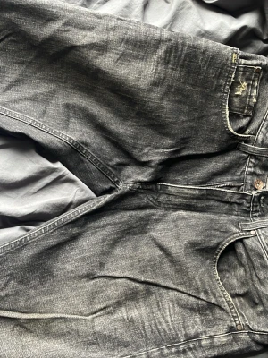 Svarta jeans med slitningar - Säljer ett par svarta jeans med snygga slitningar och tvättad look. Jeansen har klassisk femficksdesign och raka ben. Materialet är denim och färgen är svart med grå toner. Perfekt för dig som gillar en avslappnad och trendig stil.