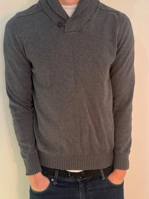 Grå stickad tröja från Selected Homme - Mörkgrå stickad tröja från Selected Homme med sjalkrage och knappdetalj vid halsen. Tröjan har långa ärmar och ribbade muddar vid ärmslut och nederkant. Perfekt för lager på lager och enkel att matcha med jeans.