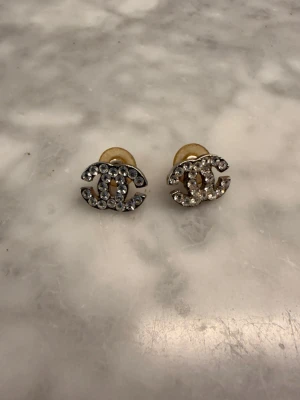Örhängen med strass CC - Säljer ett par ikoniska CC örhängen i silver metall med glittrande strass-stenar. Örhängena har den klassiska dubbla C-loggan och är perfekta för dig som vill ha något extra till din look.