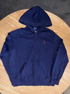 Polo Ralph Lauren Zip Hoodie Navy – Youth XL (18-20) – Fläckar - Säljer en Polo Ralph Lauren zip-hoodie i navy.  Klassisk modell med röd polo-logo på bröstet, huva och känguruficka.  Storlek: Youth XL / TG 18-20   (Ungdomsstorlek – motsvarar ungefär vuxen S i bredd men kortare i längd)  Skick: Använd.   Har två synliga fläckar: (se bilder) - En mörk fläck vid dragkedjan - En grönaktig fläck på nedre delen av ryggen (vänster sida)  Annars fin och tvättad. Prissatt efter defekterna.  Mått kan tas vid förfrågan. 