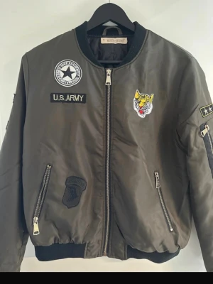 Olivgrön bomberjacka med patches - Snygg olivgrön bomberjacka med coola patches som U.S. Army, tiger och stjärnor. Jackan har dragkedjor på fickorna och ärmen, samt ribbade muddar vid ärmslut och nederkant. Materialet är glansigt och ger en klassisk streetwear-vibe.