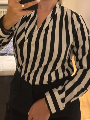 Svartvit randig blus med omlott - Snygg svartvit blus med vertikala ränder och omlottkrage. Blusen har långa ärmar med breda manschetter och en avslappnad men ändå stilren passform. Perfekt att styla med både jeans och kostymbyxor för en trendig look.