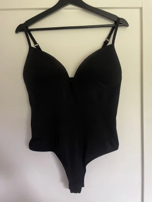 Svart body med tunna axelband - Snygg svart body med tunna, justerbara axelband och vadderade kupor. Bodyn har en djup urringning och är tillverkad i ett stretchigt material som formar sig efter kroppen. Perfekt att bära under kläder eller som ett statement-plagg.