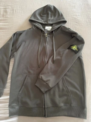 Grå hoodie från Stone Island - Säljer en grå hoodie från Stone Island med dragkedja framtill och klassisk huva med dragsko. Tröjan har kängurufickor och den ikoniska Stone Island-loggan på vänster ärm. Perfekt för dig som gillar streetwear och vill ha en stilren look.