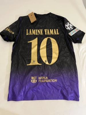 Barça matchtröja Lamine Yamal #10 - Snygg FC Barcelona matchtröja i svart och lila med guldiga detaljer och tryck. Tröjan har Lamine Yamal och nummer 10 på ryggen, samt Barça Foundation och Champions League-märke på ärmen. Tillverkad i lätt och ventilerande material, perfekt för fotboll eller träning.