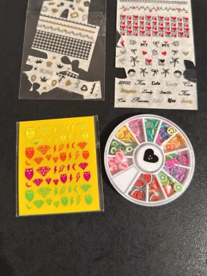 Nageldekorationer och stickers mix - Ett kit med nageldekorationer och stickers i olika färger och former. Innehåller neonfärgade stickers med motiv som diamanter, hjärtan och blixtar, samt små dekorationer i plast som frukter och pärlor i en rund ask. Perfekt för att skapa unika och kreativa naglar.