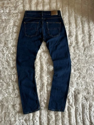 Nudie Jeans Grim Tim jeans i mörkblå - stl W31/L34 - Nudie Jeans Grim Tim i mörkblå, storlek W31/L34. 99% bomull, 1% elastan. Slim fit med rakt ben och knappgylf. Tvättnamn "New Ink".  Använd men i mycket fint skick, inga hål eller fläckar. Mått plagget platt: midjevidd 40 cm, lårvidd 25,5 cm, innerben 84,5 cm, benöppning 16 cm.  Rök- och djurfritt hem. Skriv gärna om du vill ha fler bilder eller undrar något!