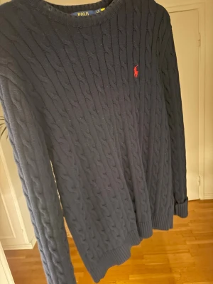 Mörkblå kabelstickad tröja Polo Ralph Lauren - Kabelstickad mörkblå tröja från Polo Ralph Lauren med rund halsringning och den klassiska röda logotypen broderad på bröstet. Tröjan har långa ärmar och ribbade muddar vid ärmslut och nederkant. Perfekt för en stilren och avslappnad look.