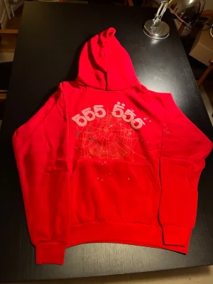 Röd Sp5der Web Hoodie – Rosa Detaljer - Röd hoodie med stort spindelnätsmotiv, rosa upphöjda detaljer och små stjärndetaljer framtill. Snygg streetwear-look med huva och känguruficka. Helt oanvänd och i nyskick. Förvarad i skyddspåse. Se bilder för detaljer.