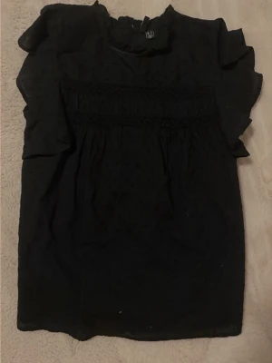 Svart blus med volangärmar från Vero Moda - Snygg svart blus med volangdetaljer på ärmarna och smockad överdel. Blusen har en lätt transparent känsla och prickigt mönster i tyget. Perfekt att styla med jeans eller kjol för en trendig look.
