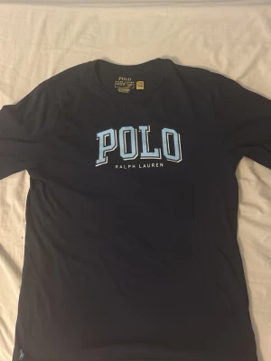  POLO Ralph Lauren långärmad - Snygg mörkblå t-shirt från POLO Ralph Lauren med stor logga i ljusblått och vitt på bröstet. Klassisk rund halsringning och korta ärmar. Perfekt för en avslappnad och stilren look.