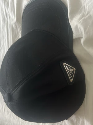 Svart keps från Prada - Snygg svart keps från Prada med klassisk böjd skärm och ikonisk metallogo på sidan. Tillverkad i ett glansigt, slitstarkt material som ger en exklusiv känsla. Perfekt accessoar för att lyfta din outfit.