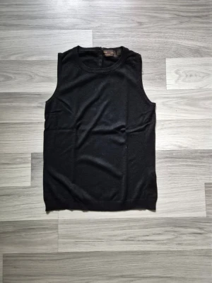 Svart ärmlös linnetopp - En stilren svart linnetopp i mjukt material. Toppen är ärmlös med rund halsringning och knäppning bak i nacken. Perfekt att matcha med jeans eller kjol för en enkel och snygg look. Sprucken i sömnen på vänster axel, är lätt att fixa för den händiga. 