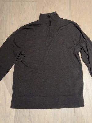 Mörkbrun tröja med halv dragkedja - Mörkbrun långärmad tröja med hög krage och halv dragkedja framtill. Tröjan har en enkel och stilren design, perfekt för lager på lager. Materialet ser ut att vara mjukt och tunt stickat, vilket gör den skön att bära.
