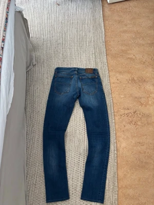 Nudie jeans - Säljer ett par mörkblå jeans med raka ben och diskreta slitningar framtill. Jeansen har klassisk femficksdesign och bruna detaljer vid linningen. Perfekta till en avslappnad stil och passar till det mesta i garderoben.