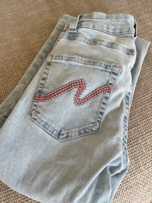Nelly - Ljusblå jeans med brodyr - Säljer ett par ljusblå jeans med raka ben och röda broderade detaljer på bakfickorna. Jeansen har klassisk femficksdesign och är gjorda i mjukt denimtyg. 