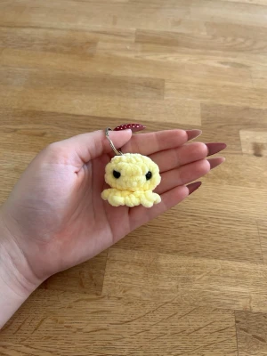 Gul virkad bläckfisk nyckelring - Söt handgjord nyckelring i form av en liten gul bläckfisk. Tillverkad i mjukt garn med svarta virkade ögon. Perfekt att hänga på väskan eller nyckelknippan för en personlig touch.