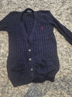 Mörkblå kabelstickad kofta Polo Ralph Lauren - Kabelstickad mörkblå kofta från Polo Ralph Lauren med knappar framtill, två fickor och röd broderad logga på bröstet. Långärmad modell med ribbade muddar och v-ringad hals. Perfekt för lager på lager och klassisk preppy stil.