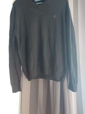 Svart stickad tröja från Ralph Lauren - Klassisk svart stickad tröja från Ralph Lauren med V-ringning och den ikoniska gröna loggan broderad på bröstet. Tröjan har långa ärmar och ribbade muddar vid ärmslut och nederkant. Perfekt till lager på lager och enkel att matcha.