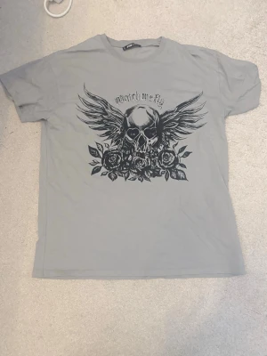 Grå t-shirt med döskalletryck - Säljer en grå t-shirt med ett stort svart tryck av en döskalle med vingar och rosor på framsidan. Texten 'Watch Me Fly' ovanför motivet. Klassisk passform och korta ärmar, perfekt för dig som gillar edgy stil.