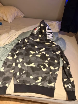  Bape Camo hoodie med hajmotiv och dragkedja - Säljer en bape camouflagemönstrad hoodie med hajmotiv på huvan och dragkedja framtill. Tröjan har svarta muddar vid ärmslut och midja samt vita och grå detaljer. På ena ärmen finns en rund logga. Perfekt för dig som vill sticka ut med streetwear-stil. Säljer den pågrund av att den är för liten för mig. Den är knappt använd.