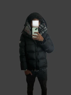 Burberry jacka  - Snygg och stilren Burberry vinterjacka i svart. Varm och perfekt för höst/vinter. Klassisk Burberry-design med fodrad huva och en clean look som passar till det mesta.