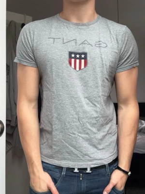 Grå GANT t-shirt med  - Säljer en grå t-shirt från GANT med ett tryck av deras klassiska sköld med stjärnor och ränder på bröstet. T-shirten har rund halsringning och korta ärmar. Perfekt till jeans för en clean och enkel look.