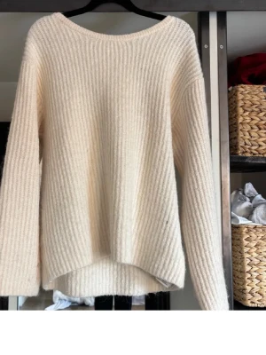 Krämvit stickad tröja med rosett - Supersöt krämvit stickad tröja med ribbad struktur och lång ärm. Tröjan har en snygg v-ringning i ryggen och en stor dekorativ rosett upptill. Perfekt för dig som gillar en mjuk och stilren look.
