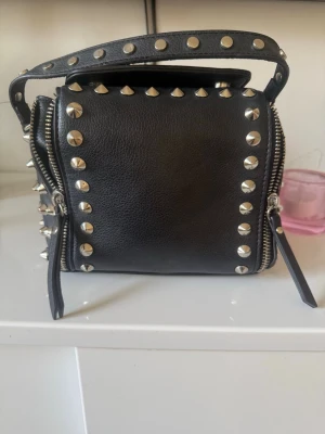 Handväska zara - Cool svart handväska i fyrkantig modell med silverfärgade nitar runt kanterna och på handtaget. Väskan har dragkedjor på sidorna och är gjord i ett läderliknande material. Perfekt för dig som gillar en edgy stil och vill sticka ut.