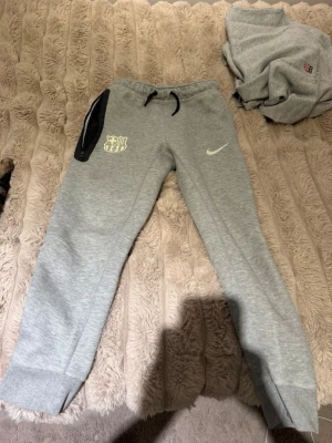 Gråa Nike FCB mjukisbyxor - Snygga grå mjukisbyxor från Nike med FC Barcelona-logga på vänster ben och svart dragkedja vid fickan. Byxorna har snörning i midjan och är perfekta för chill eller träning. Sköna och stilrena med en loose passform. Tröjan finns även inne på profilen 