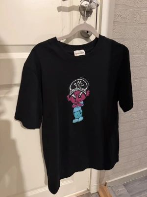 Svart Moncler t-shirt med tryck - Svart t-shirt från Moncler med ett färgglatt tecknat tryck framtill. T-shirten har rund hals och korta ärmar. Perfekt för dig som vill ha en cool och unik look med streetwear-vibe.