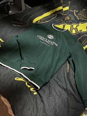 Grön sweatshirt Legacy Manor - Mörkgrön sweatshirt storlek med vit text och tryck på bröstet där det står 'Legacy Manor Health & Wellness Club'. Tröjan har vita detaljer vid krage och nederkant samt långa ärmar. Perfekt för en avslappnad och stilren look. Från Pier-one Collection 