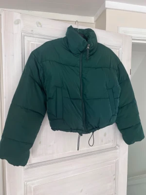 Mörkgrön cropped streetstyle pufferjacka - Mörkgrön pufferjacka från H&M i storlek XS. Kort och trendig modell med hög krage, dragkedja och justerbar dragsko nedtill. Perfekt till de kallare dagarna framöver. Stor i storleken, passar bra även till S &M. Mycket fint skick utan anmärkningar. Använd 2 gånger.