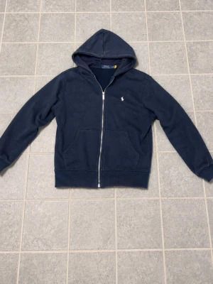 Mörkblå hoodie från Polo Ralph Lauren - Snygg mörkblå hoodie från Polo Ralph Lauren med dragkedja framtill och klassisk huva. Tröjan har två fickor på magen och den ikoniska lilla loggan broderad på bröstet. Perfekt för en avslappnad och stilren look, den är liten i storlek så den passar som S.