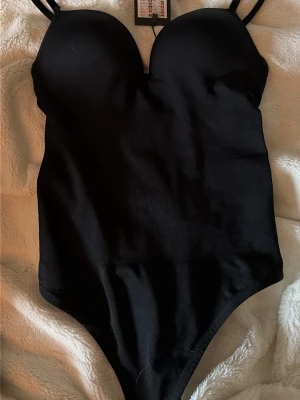 Svart body med vadderade kupor - Snygg svart body med tunna justerbara axelband och vadderade kupor för extra stöd. Tillverkad i stretchigt material som sitter tight mot kroppen och ger en smickrande siluett. Perfekt att styla under skjortor eller tröjor.