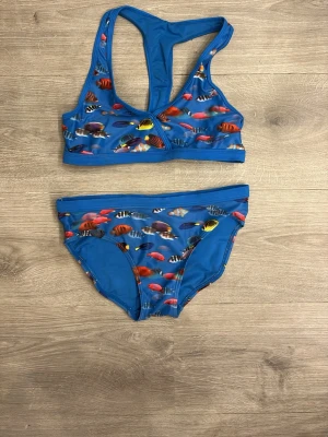Blå bikini med fiskmönster - Säljer en blå bikini med färgglatt fiskmönster. Överdelen har sportig modell med breda axelband och underdelen är klassisk med medelhög midja. Materialet är stretchigt och snabbtorkande, perfekt för stranden eller poolen.