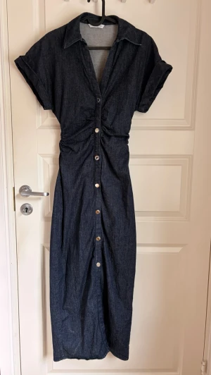 Mörkblå jeansklänning med knappar - Snygg mörkblå långklänning i jeansmaterial med korta ärmar, krage och knappar längs hela framsidan. Klänningen har en markerad midja och en avslappnad passform. Perfekt för dig som gillar en stilren och tidlös look.