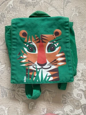 Grön ryggsäck från Tiger ryggsäck från Coq en Pâte.  - Säljer en grön ryggsäck i tyg med ett coolt tigeransikte tryckt på framsidan. Ryggsäcken har justerbara axelband och ett praktiskt handtag upptill. Perfekt för dig som vill sticka ut med en färgglad och lekfull design.