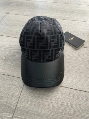 Fendi keps - Snygg svart keps från Fendi med klassiskt FF-monogram i tyg på ovandelen och stilren skärm i svart läder. Säljs då den aldrig kommit till användning och försent för att lämna tillbaka.