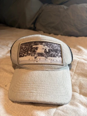 Grå och svart keps med fotomotiv - Säljer en unik keps i grått och svart med mesh baktill och justerbar snapback. Framtill finns ett svartvitt fotomotiv av en fotbollsspelare i action. Kepsen har böjd skärm och är tillverkad i mjukt tyg framtill och mesh bak.
