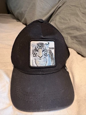 Svart truckerkeps med tigerpatch - Säljer en svart truckerkeps med mesh på sidorna och ett broderat tygmärke framtill med en tiger. Kepsen har böjd skärm och justerbar snapback baktill. Perfekt för dig som vill ha en cool och sportig look.