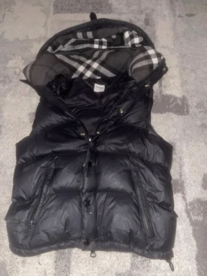 Svart dunväst från Burberry - Snygg svart dunväst från Burberry med klassiskt rutigt foder i huvan. Västen har dragkedja och knappar framtill samt två dragkedjeförsedda fickor. Perfekt för lager på lager och ger en stilren look med exklusiv känsla. Pris kan diskuteras vid snabb affär! Armarna kommer inte med. 