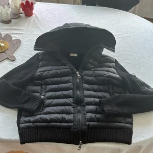 Svart Moncler pufferjacka med huva - Svart pufferjacka från Moncler med stickade ärmar och ribbade muddar. Jackan har quiltad front och huva, dragkedja framtill och en Moncler-logga på ärmen. Materialmix av polyester och stickat ger en snygg och fräsch look. Jackan är ca 1 år gammal och inte använd allt för mycket. Sjukt bra kvalite. Skriv till mig om ni vill ha fler bilder alltid redo o svara👍🏻👍🏻