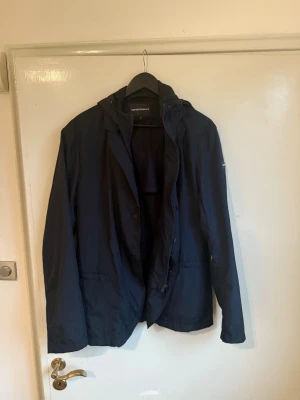Armani höst jacka - Snygg mörkblå vindjacka från Giorgio Armani med huva och dragkedja framtill. Jackan har en minimalistisk design, långa ärmar och praktiska fickor. Perfekt för blåsiga dagar och enkel att matcha med olika outfits.