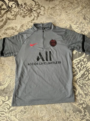 Grå PSG träningströja från Nike - Snygg grå träningströja från PSG med halv dragkedja, svarta detaljer och röd Nike-logga. Tröjan har PSG-emblem på bröstet och texten 'Accor Live Limitless' framtill samt 'Visit Rwanda' på ryggen. Tillverkad i lätt och funktionellt material, perfekt för fotbollsträning.