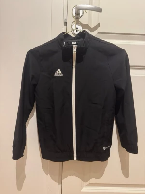 Svart träningsjacka från Adidas stl 128 - Snygg svart träningsjacka från Adidas med vit dragkedja och logga på bröstet. Jackan har lång ärm, hög krage och elastisk nederkant. Tillverkad av återvunnet material och passar perfekt till träning eller sportaktiviteter.