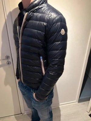 Moncler daniel dunjacka  - Snygg svart dunjacka från Moncler med quiltad design och ståkrage. Jackan har dragkedja framtill med rödvita detaljer och två fickor med dragkedja. Moncler-logga på vänster ärm. Perfekt för kalla vinterdagar.